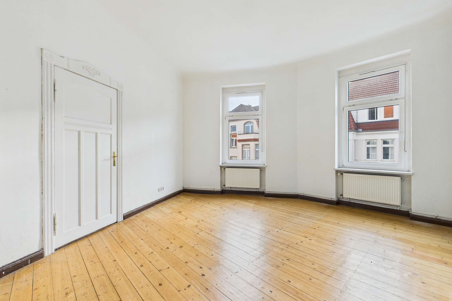 Leere, helle Wohnung mit Holzfußboden, zwei Fenstern und einer weißen Tür. Wände sind neutral gestrichen.
