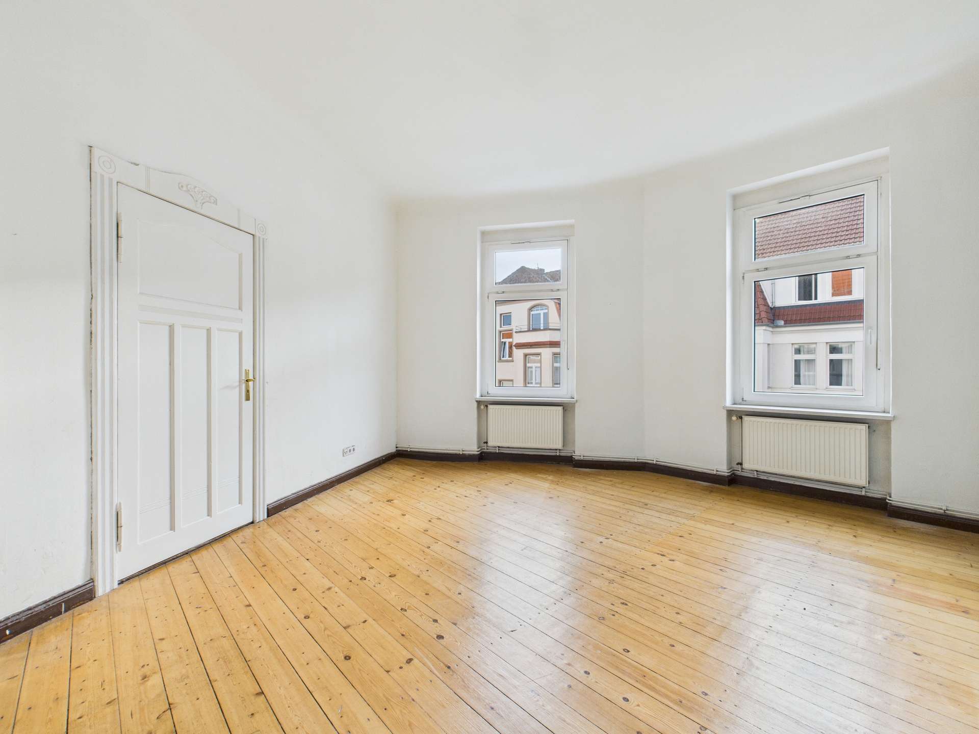 Leere, helle Wohnung mit Holzfußboden, zwei Fenstern und einer weißen Tür. Wände sind neutral gestrichen.