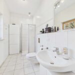 Modernes Badezimmer mit Dusche, Waschbecken, WC und großen Fenstern. Helle Fliesen und klare Linien dominieren den Raum.