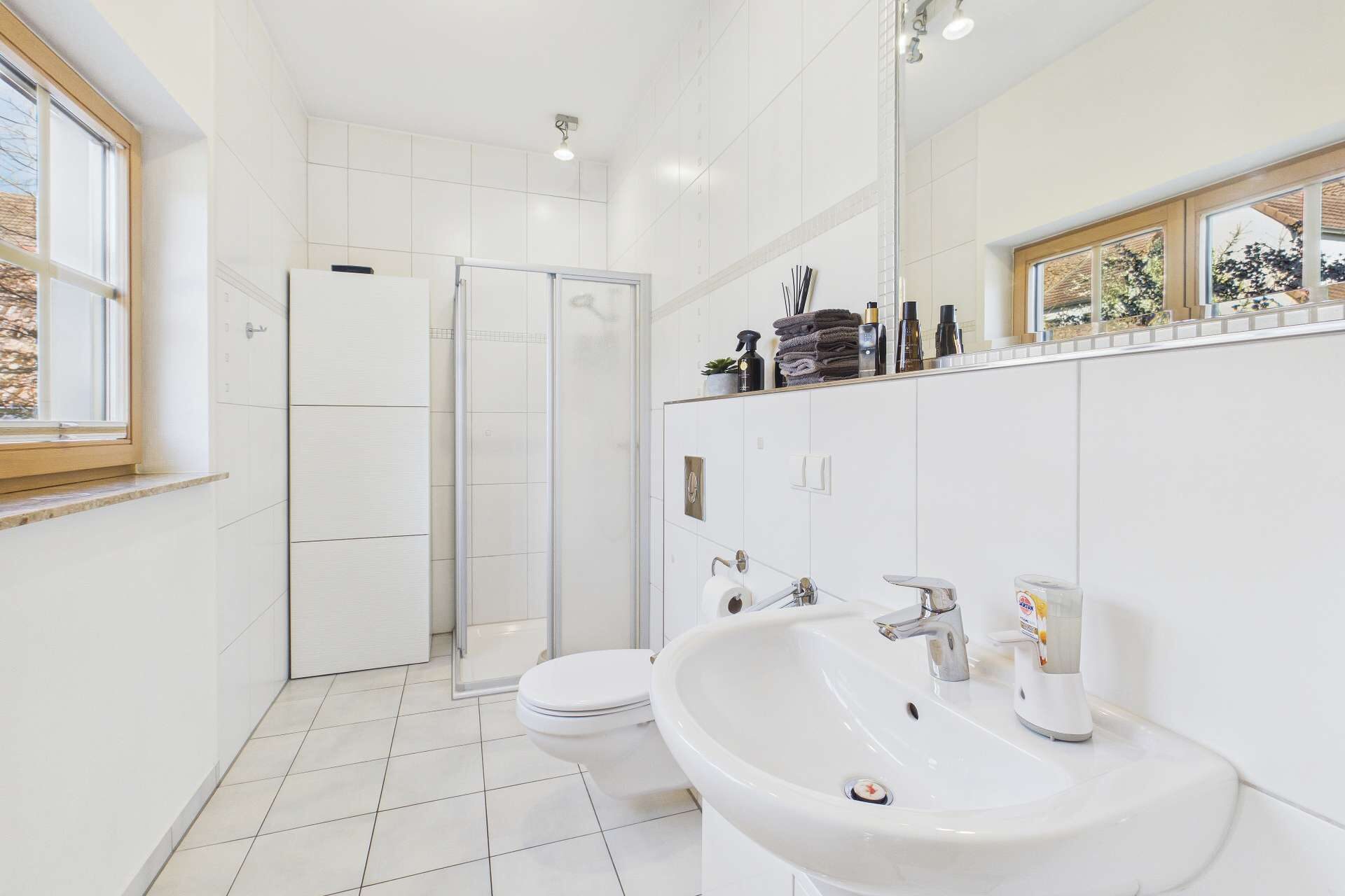 Modernes Badezimmer mit Dusche, Waschbecken, WC und großen Fenstern. Helle Fliesen und klare Linien dominieren den Raum.