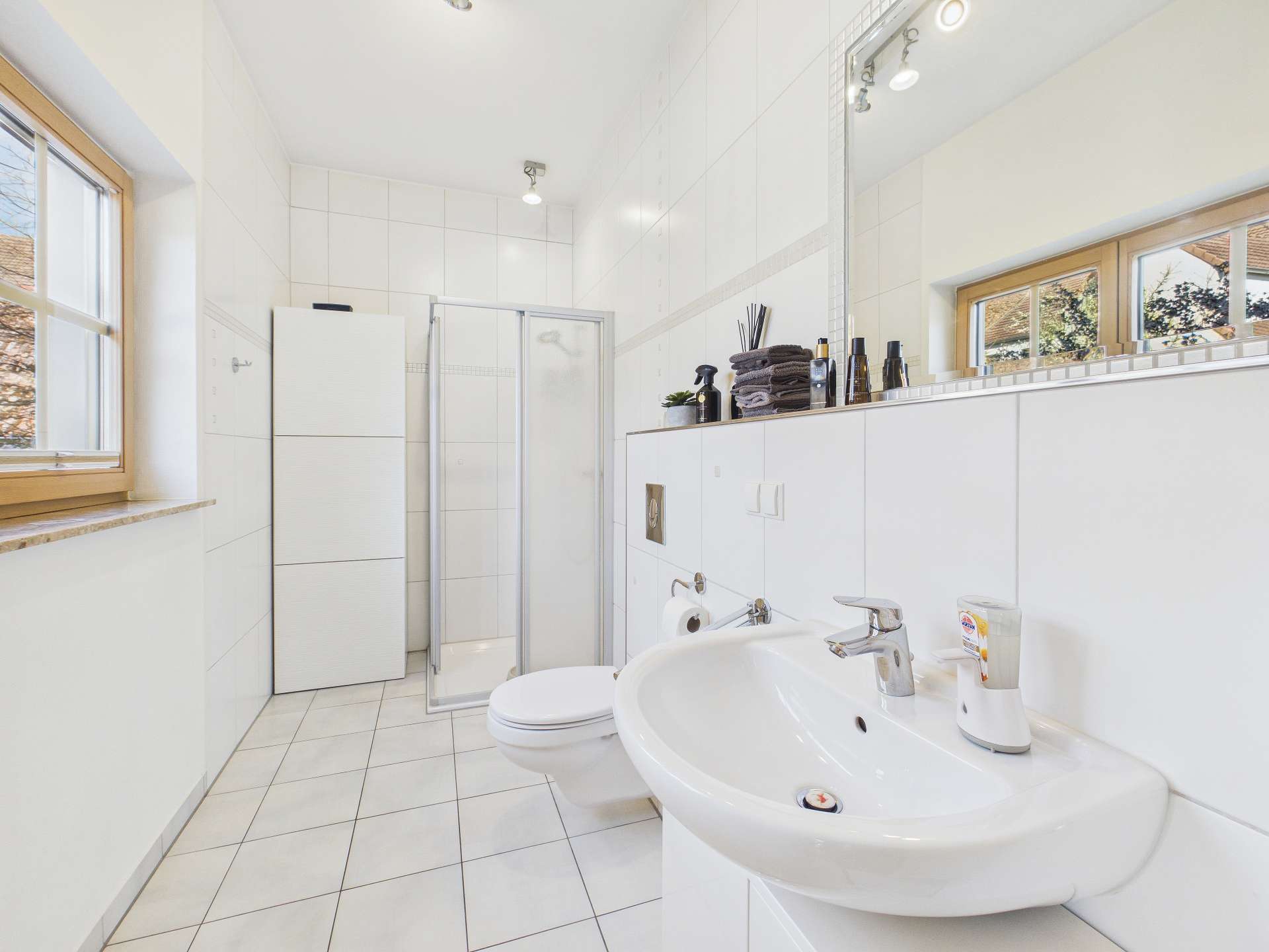 Modernes Badezimmer mit Dusche, Waschbecken, WC und großen Fenstern. Helle Fliesen und klare Linien dominieren den Raum.