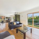 Helles Wohnzimmer mit Holzboden, schwarzem Sofa, Glastisch und großen Fenstern mit Blick auf den Garten.