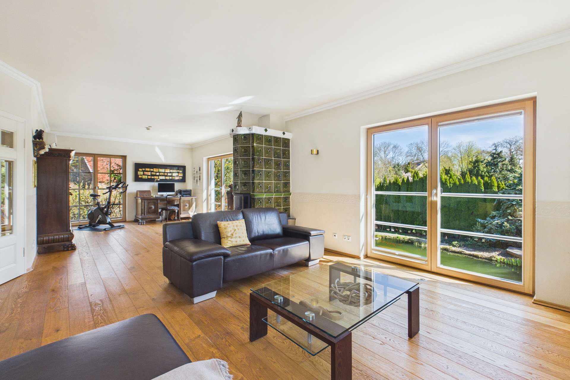 Helles Wohnzimmer mit Holzboden, schwarzem Sofa, Glastisch und großen Fenstern mit Blick auf den Garten.