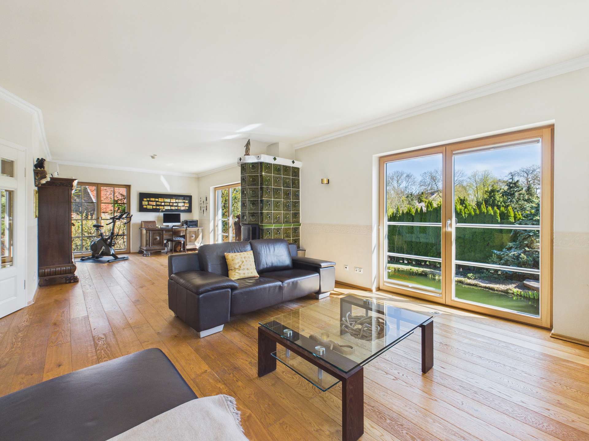Helles Wohnzimmer mit Holzboden, schwarzem Sofa, Glastisch und großen Fenstern mit Blick auf den Garten.