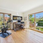 Helles Arbeitszimmer mit Schreibtisch, Computer, Fitnessgerät und großen Fenstern mit Blick ins Grüne.