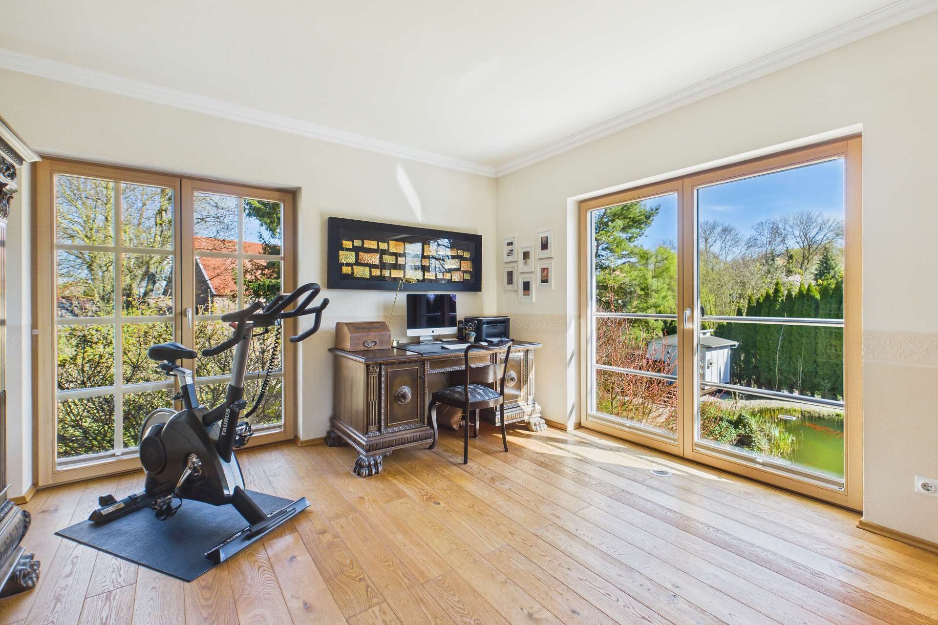 Helles Arbeitszimmer mit Schreibtisch, Computer, Fitnessgerät und großen Fenstern mit Blick ins Grüne.