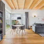 Modernes Wohnzimmer mit Holzdecke, grauem Sofa, runder Esstisch und Bücherregal. Großer Fensterblick auf den Garten.