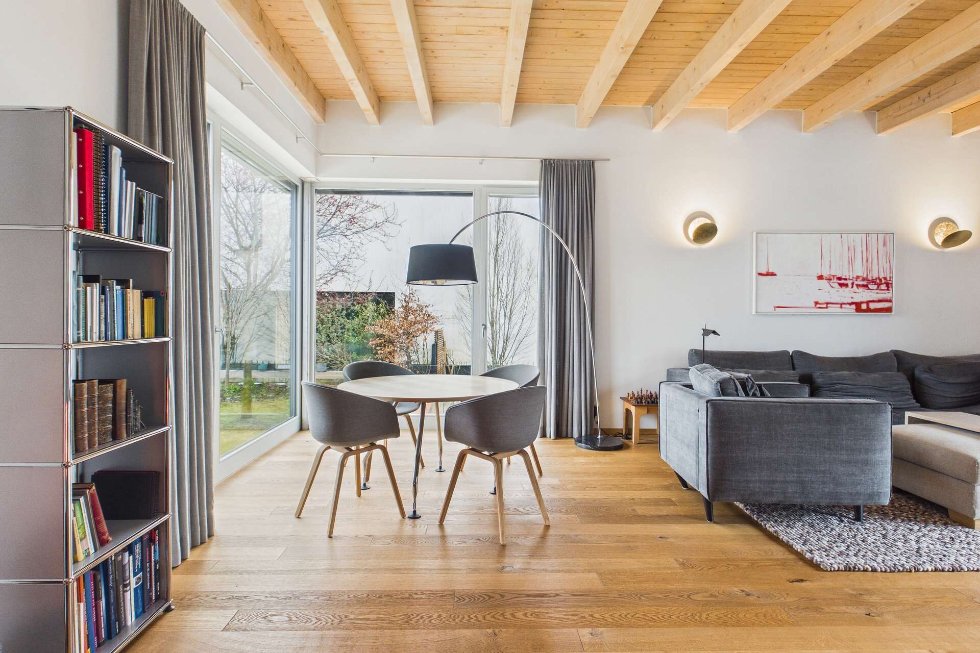 Modernes Wohnzimmer mit Holzdecke, grauem Sofa, runder Esstisch und Bücherregal. Großer Fensterblick auf den Garten.
