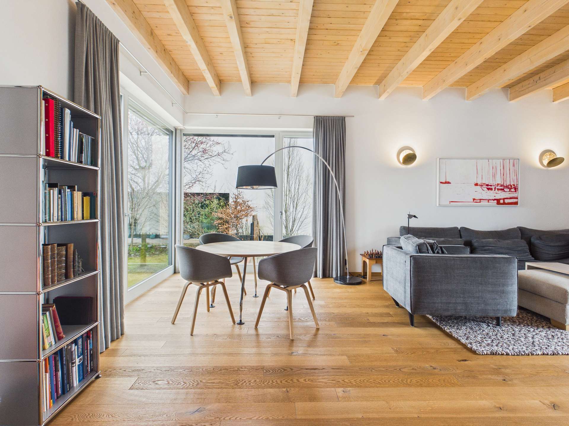 Modernes Wohnzimmer mit Holzdecke, grauem Sofa, runder Esstisch und Bücherregal. Großer Fensterblick auf den Garten.