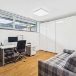 Modernes Arbeitszimmer mit Schreibtisch, Computer, Bürostuhl und Couch, helles Licht durch große Fenster.