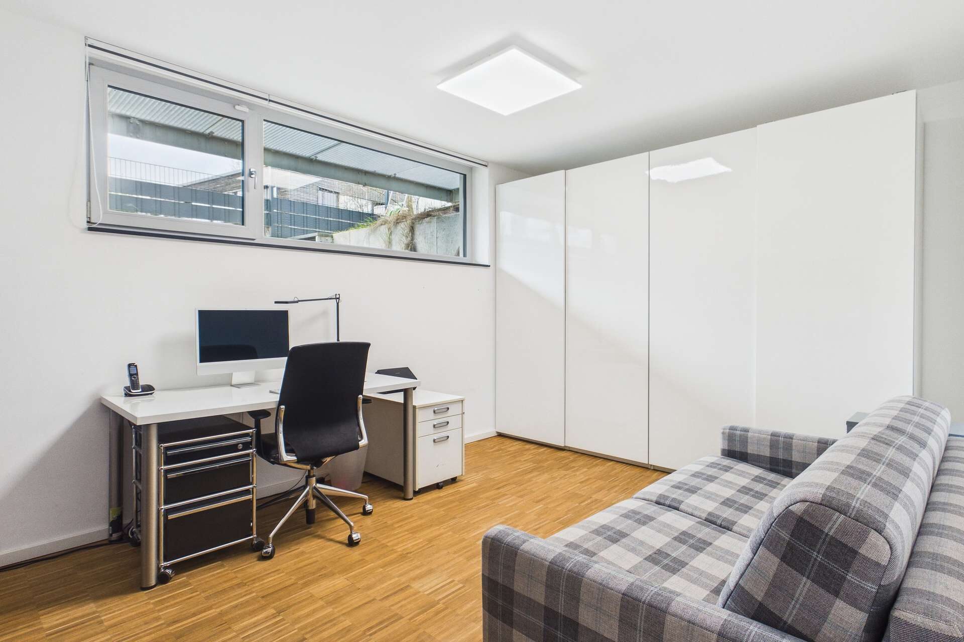 Modernes Arbeitszimmer mit Schreibtisch, Computer, Bürostuhl und Couch, helles Licht durch große Fenster.