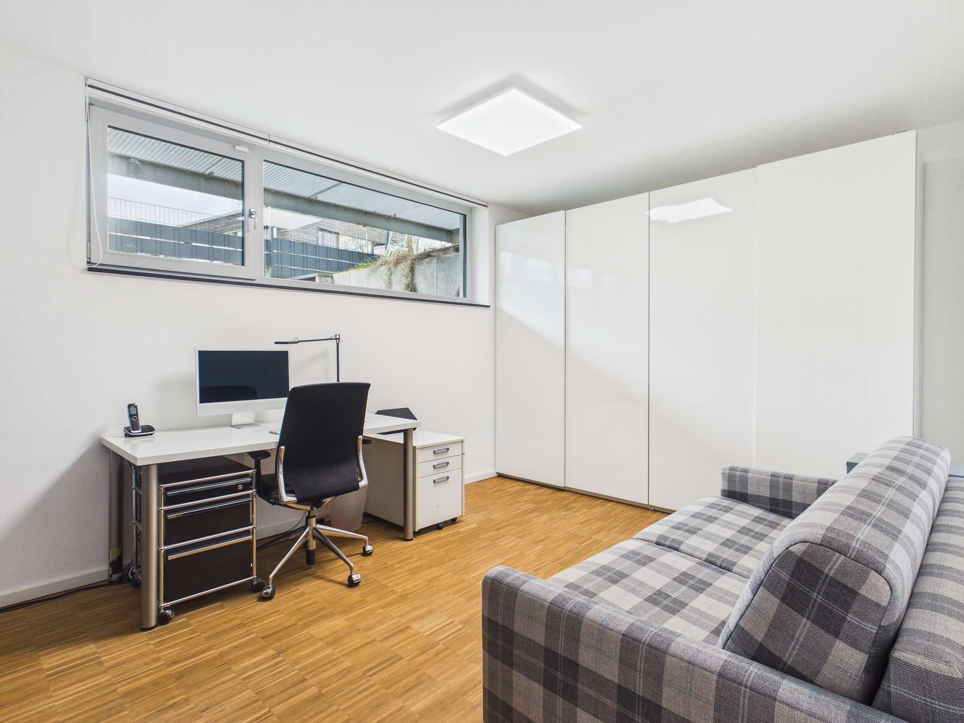 Modernes Arbeitszimmer mit Schreibtisch, Computer, Bürostuhl und Couch, helles Licht durch große Fenster.