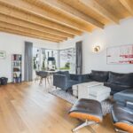 Modernes Wohnzimmer mit Holzdecke, gemütlichem Sofa, Kamin und großem Fenster, das auf eine Landschaft blickt.