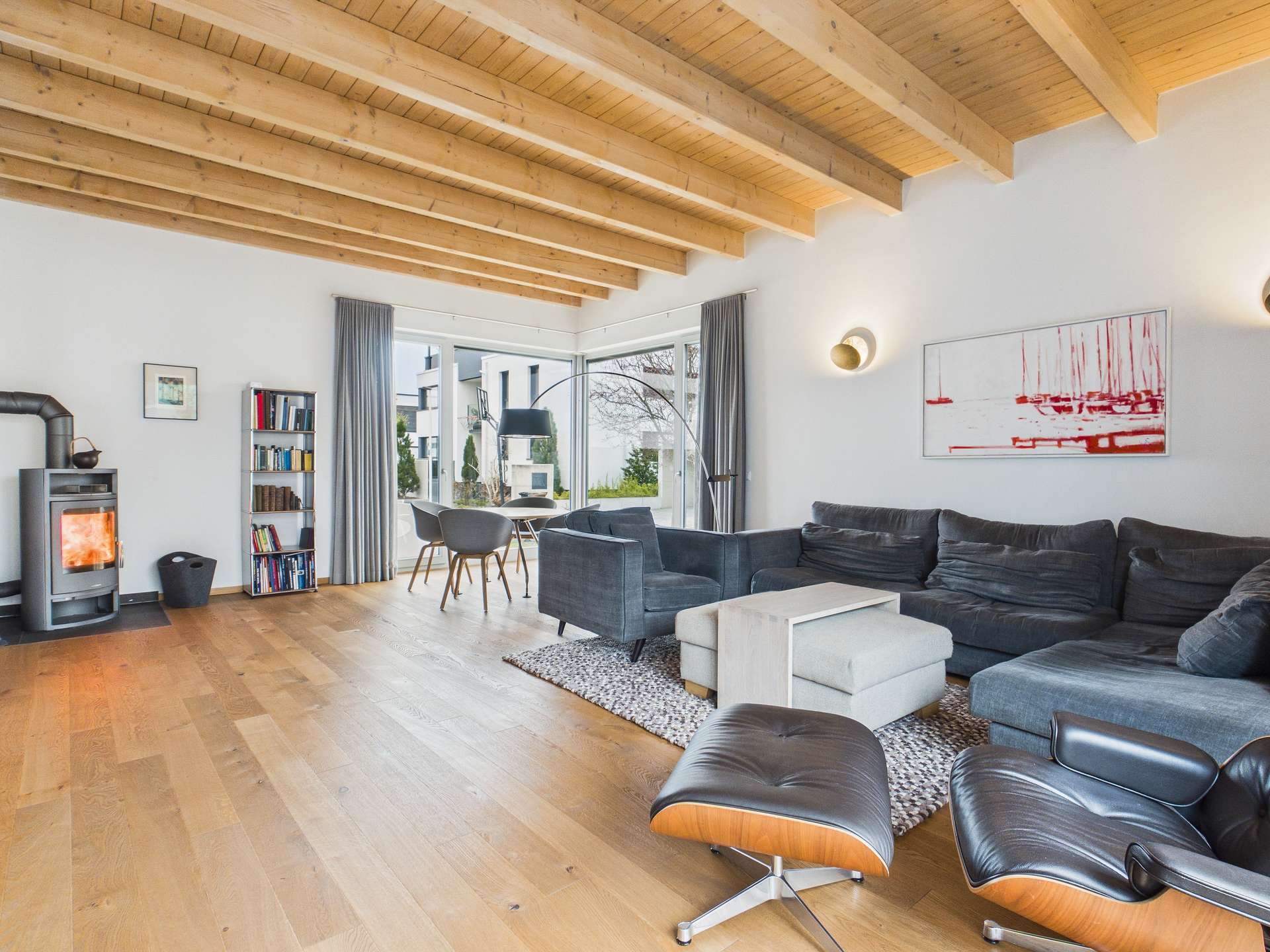 Modernes Wohnzimmer mit Holzdecke, gemütlichem Sofa, Kamin und großem Fenster, das auf eine Landschaft blickt.