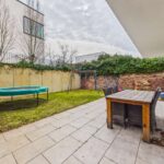 Gartenbereich mit Rasen, Trampolin, Schaukel und Essgruppe aus Holz. Mauer im Hintergrund, grauer Himmel.