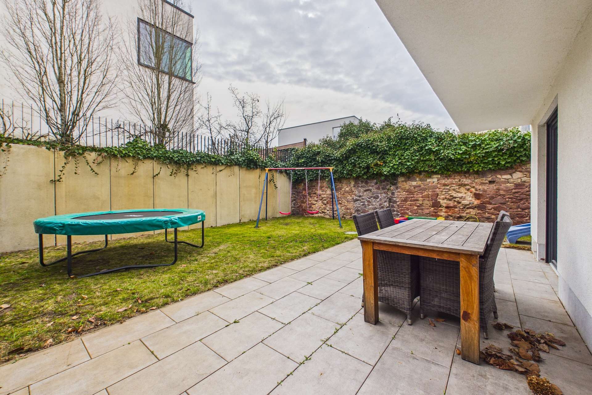 Gartenbereich mit Rasen, Trampolin, Schaukel und Essgruppe aus Holz. Mauer im Hintergrund, grauer Himmel.