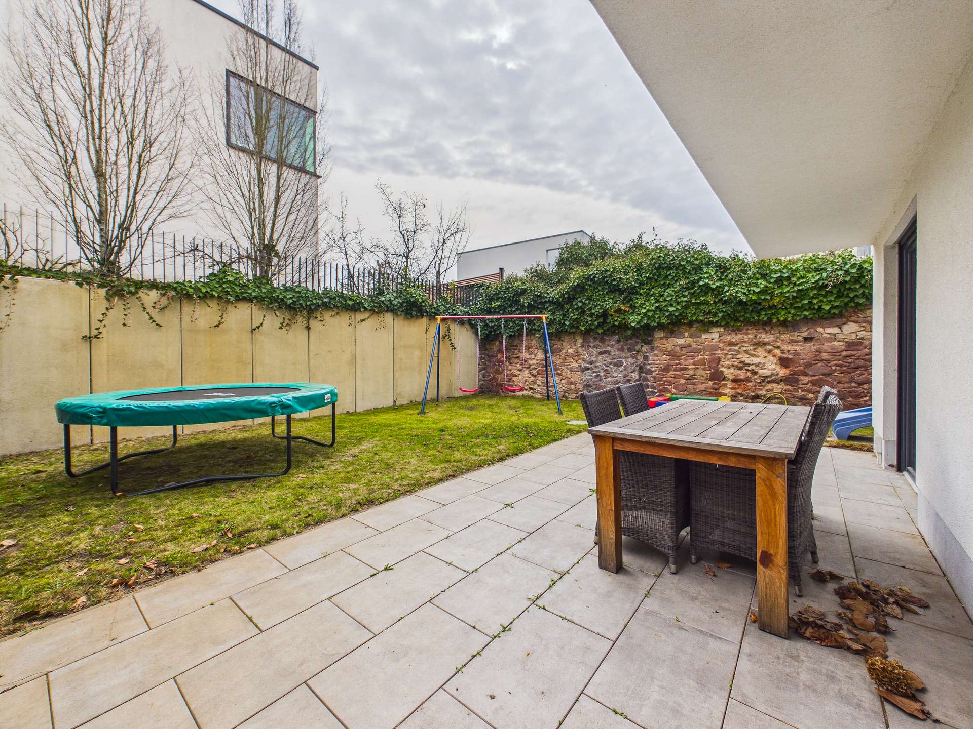 Gartenbereich mit Rasen, Trampolin, Schaukel und Essgruppe aus Holz. Mauer im Hintergrund, grauer Himmel.