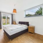Schlafzimmer mit Doppelbett, Holzboden, Fenster mit Blick auf Garten und heller Beleuchtung.