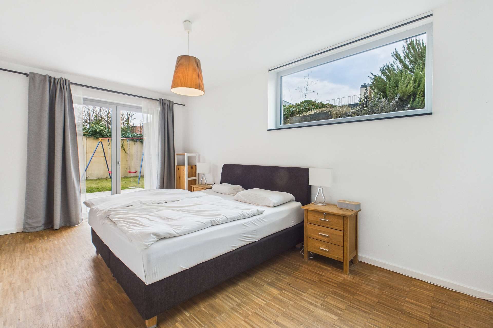 Schlafzimmer mit Doppelbett, Holzboden, Fenster mit Blick auf Garten und heller Beleuchtung.