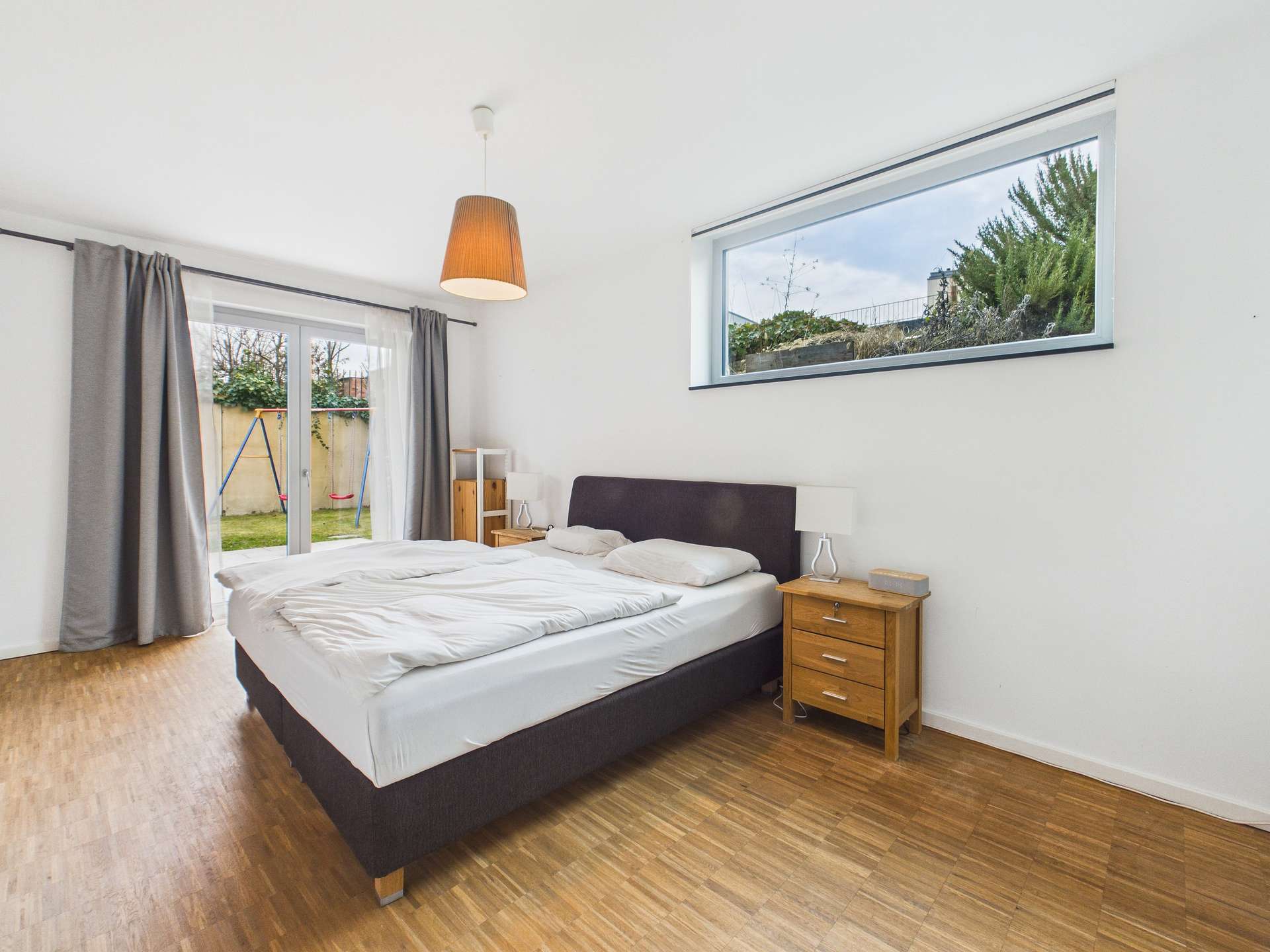 Schlafzimmer mit Doppelbett, Holzboden, Fenster mit Blick auf Garten und heller Beleuchtung.