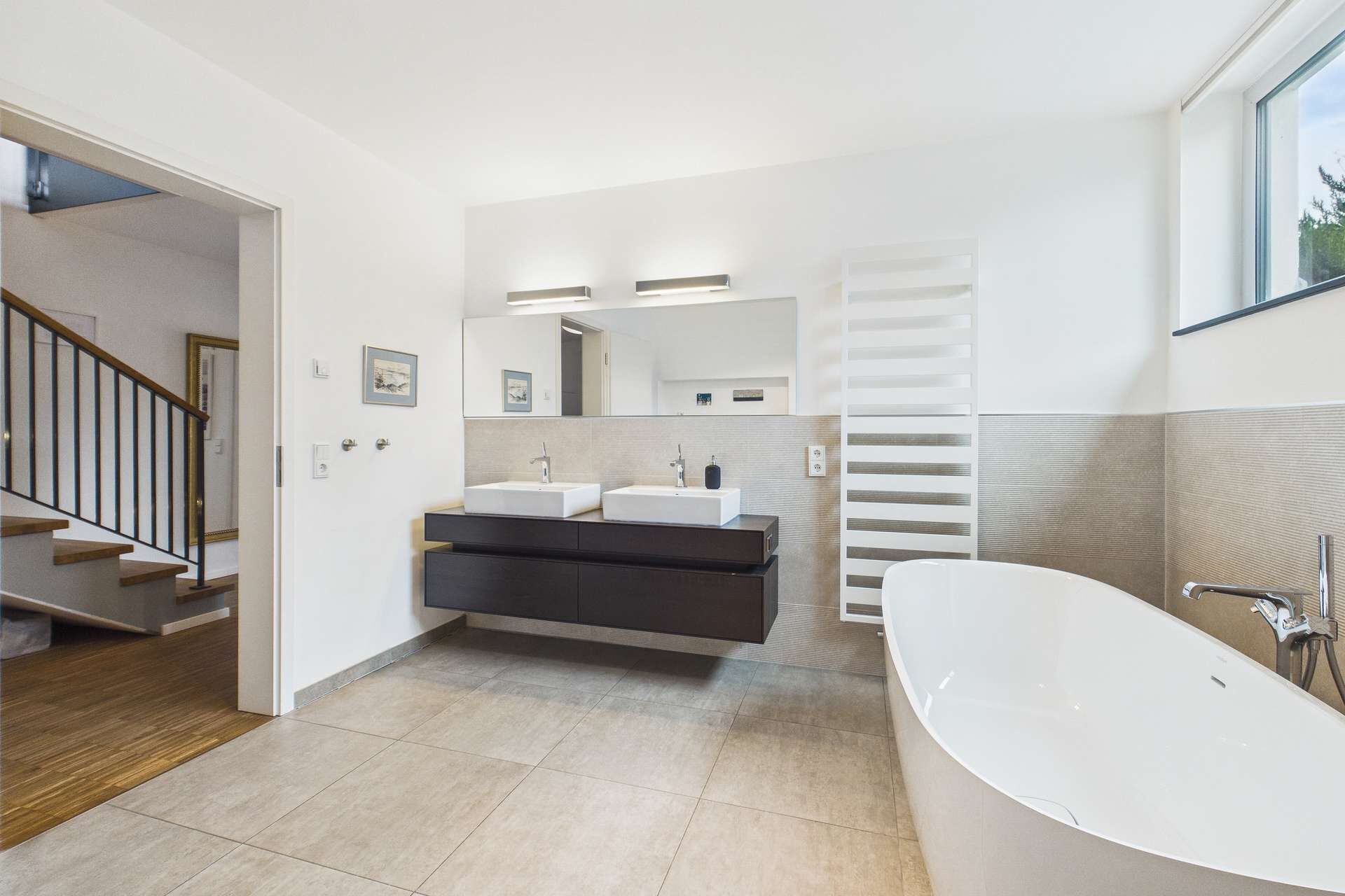 Modernes Badezimmer mit freistehender Badewanne, Doppelwaschbecken und Holztreppe im Hintergrund.