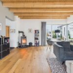 Helles Wohnzimmer mit Holzdecke, Kamin, grauem Sofa und Essbereich, große Fenster mit Blick ins Freie.