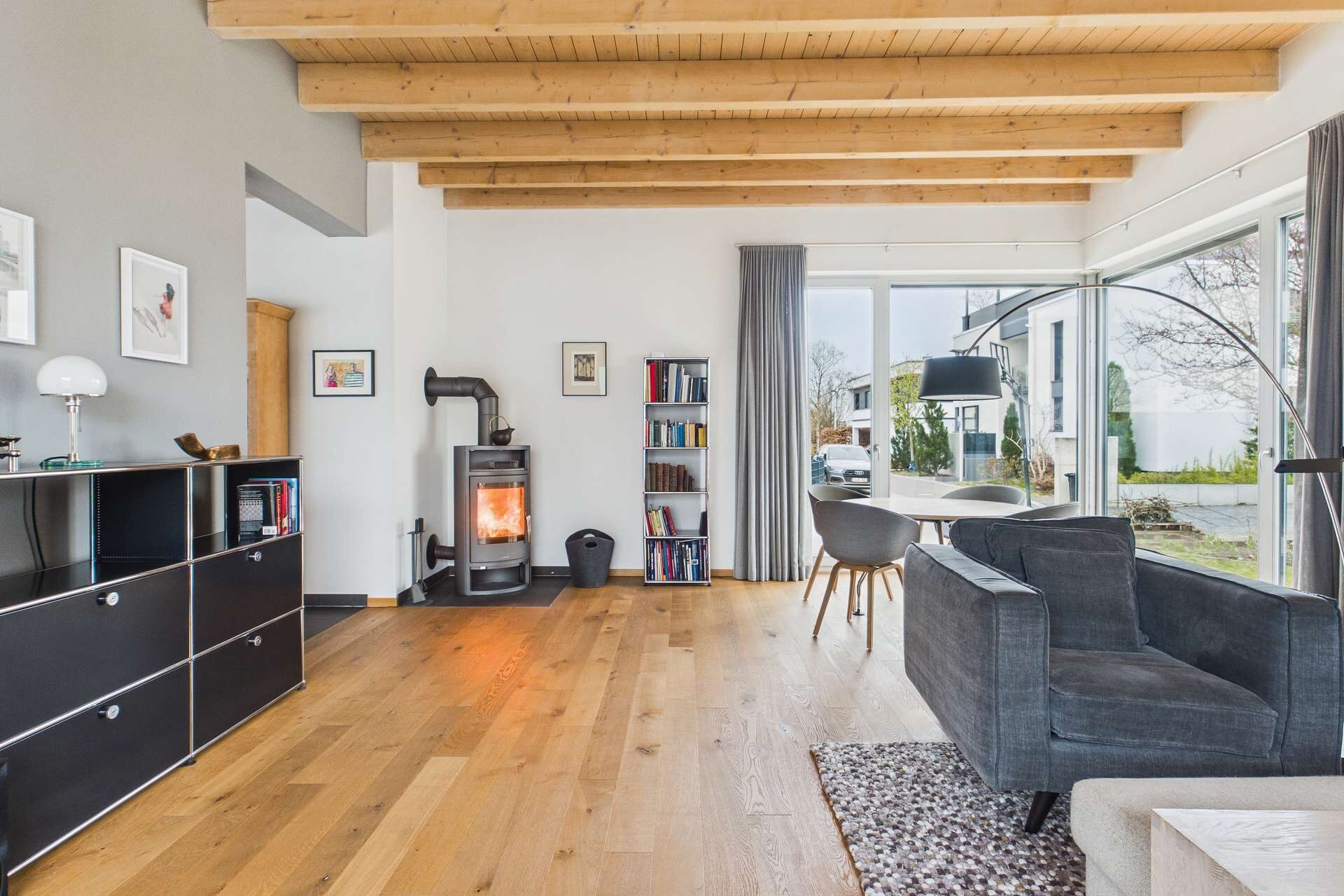 Helles Wohnzimmer mit Holzdecke, Kamin, grauem Sofa und Essbereich, große Fenster mit Blick ins Freie.