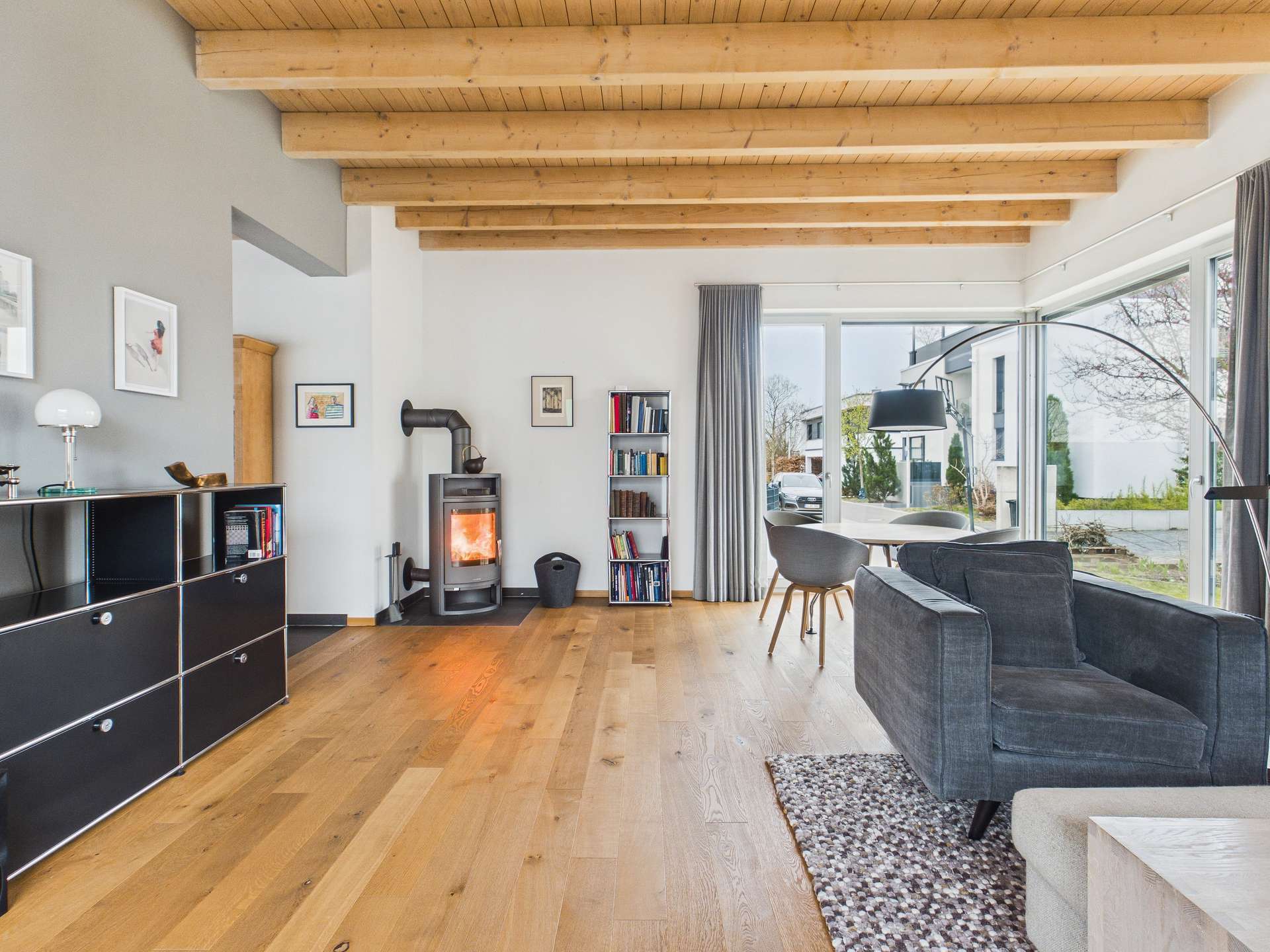 Helles Wohnzimmer mit Holzdecke, Kamin, grauem Sofa und Essbereich, große Fenster mit Blick ins Freie.