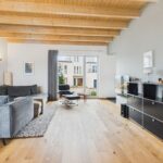 Heller Wohnraum mit Holzdecke, grauem Sofa, schwarzem Regal und großen Fenstern, die auf einen Innenhof blicken.