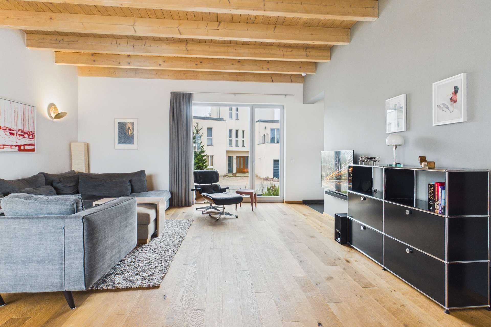Heller Wohnraum mit Holzdecke, grauem Sofa, schwarzem Regal und großen Fenstern, die auf einen Innenhof blicken.