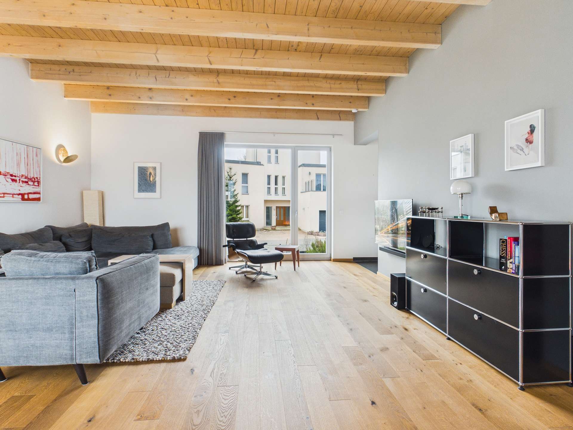 Heller Wohnraum mit Holzdecke, grauem Sofa, schwarzem Regal und großen Fenstern, die auf einen Innenhof blicken.