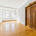 Helle, leere Wohnung mit Holzboden und großen Fenstern; eine elegante Holztür im Hintergrund.