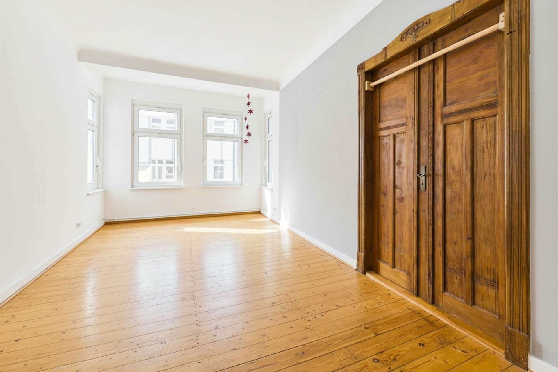 Helle, leere Wohnung mit Holzboden und großen Fenstern; eine elegante Holztür im Hintergrund.