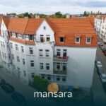 Mehrstöckiges Wohnhaus mit rotem Ziegeldach und Balkonen, umgeben von Straßen und weiteren Gebäuden.