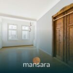 Heller Raum mit Holzfußboden und einer klassischen Holztür, weiße Wände und große Fenster.
