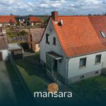 Ein zweigeschossiges Haus mit rotem Satteldach und Garten, umgeben von weiteren Gebäuden und einem klaren Himmel.
