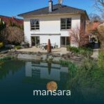Moderne Villa mit Garten und Teich, umgeben von Bäumen und Sträuchern, unter blauem Himmel.
