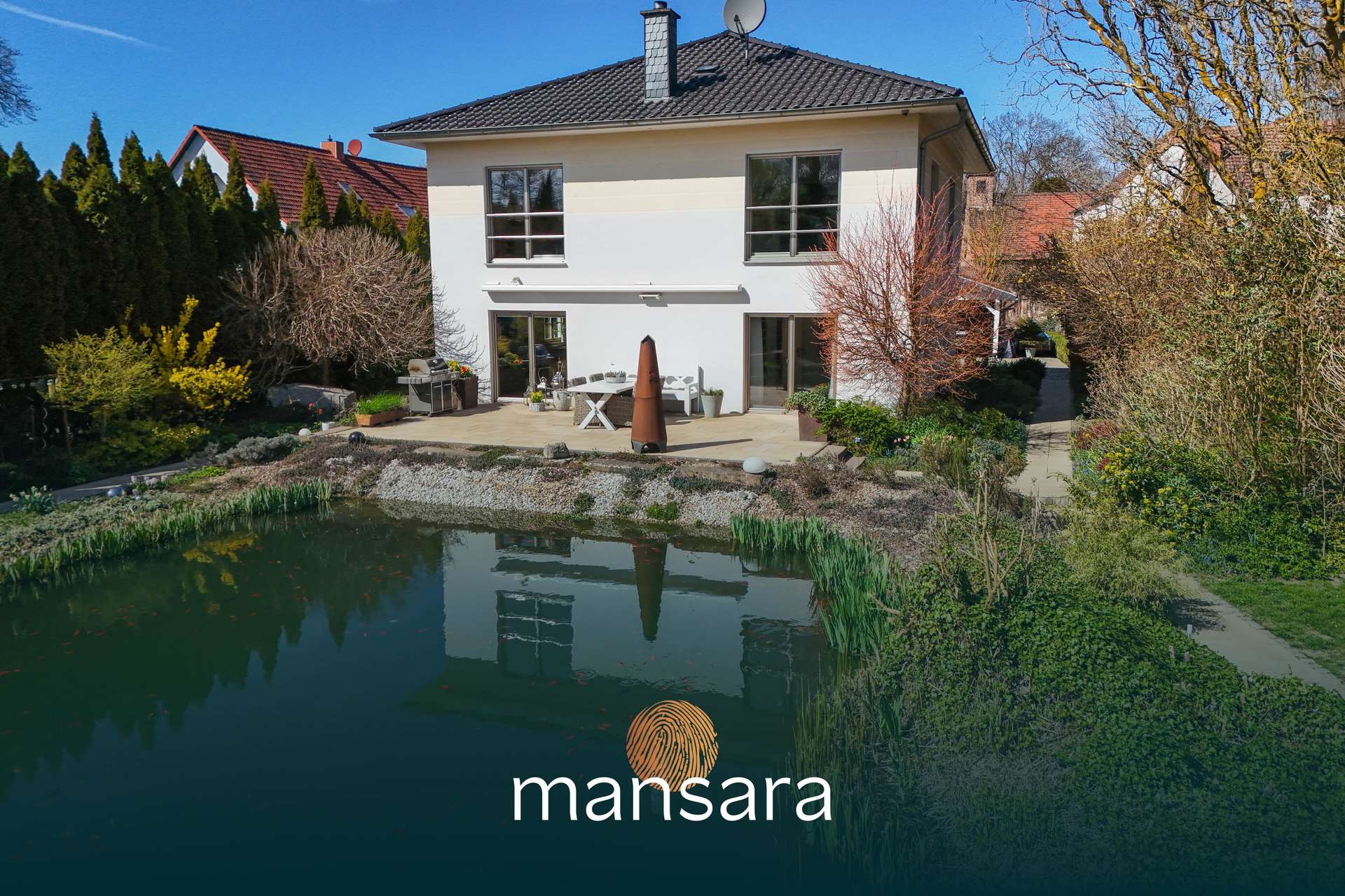 Moderne Villa mit Garten und Teich, umgeben von Bäumen und Sträuchern, unter blauem Himmel.