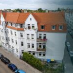 Blick auf ein mehrstöckiges Wohngebäude mit roten Dächern und bepflanzten Balkonen, umgeben von einer ruhigen Straße.