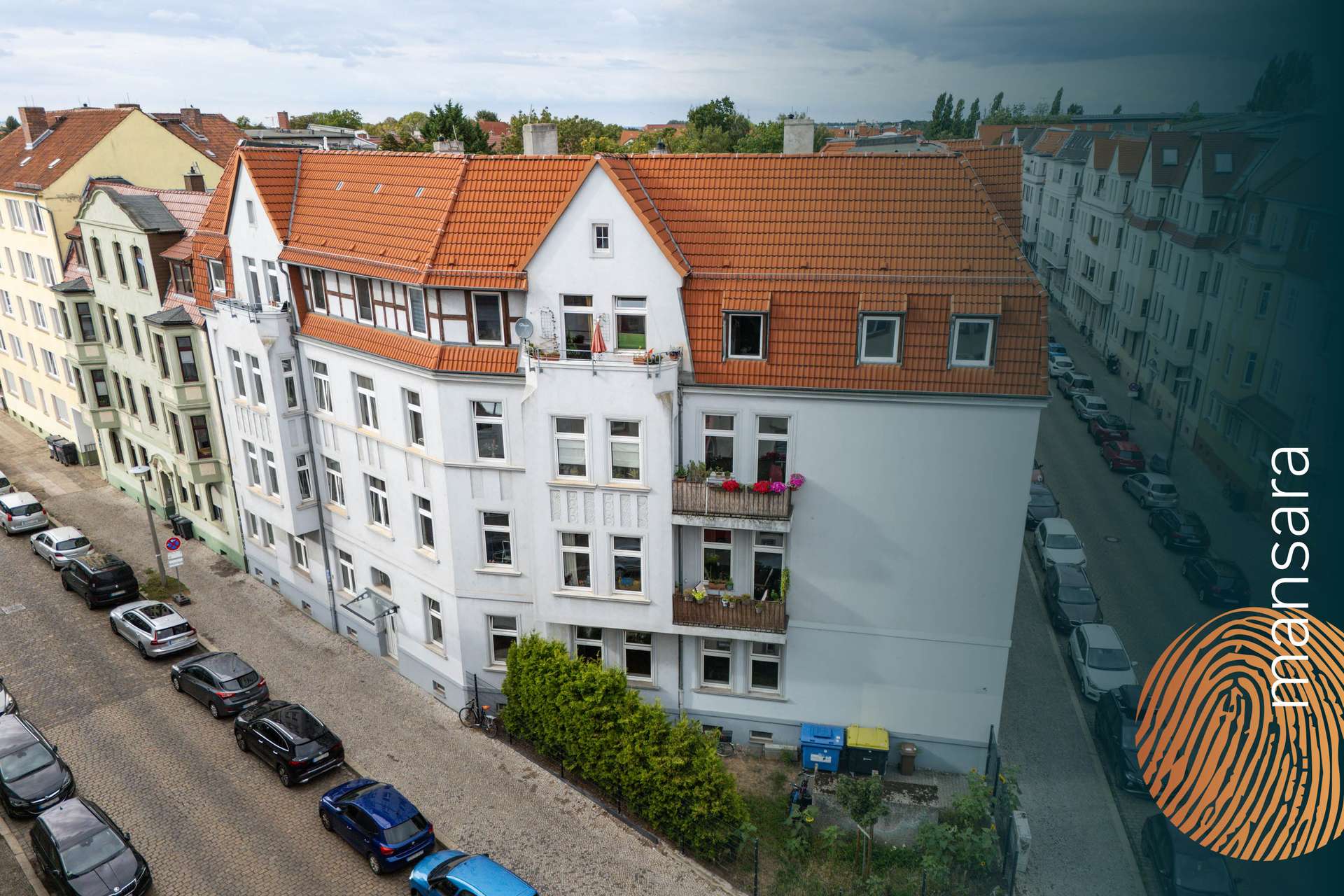Blick auf ein mehrstöckiges Wohngebäude mit roten Dächern und bepflanzten Balkonen, umgeben von einer ruhigen Straße.