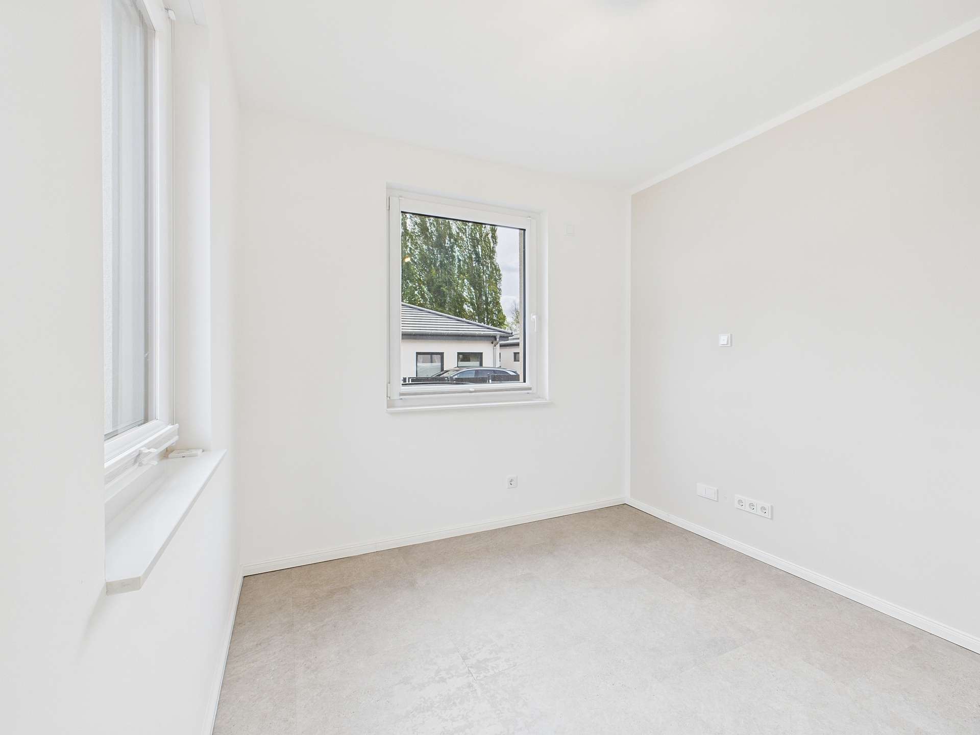 Helles, leeres Zimmer mit Fenster und neutralen Wänden, Teppichboden und Blick auf die Außenansicht.