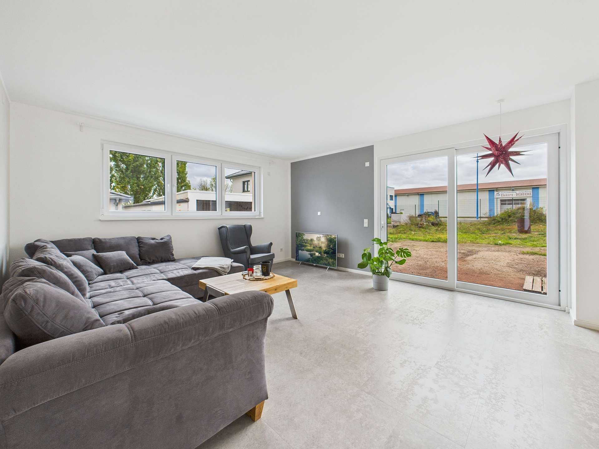 Moderner Wohnraum mit grauem Sofa, schwarzem Sessel, TV und großen Fenstern, die auf einen Außenbereich blicken.