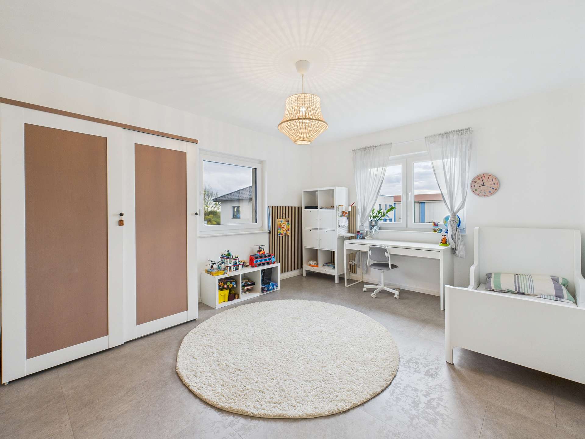 Helles Kinderzimmer mit Teppich, Schreibtisch, Regal und Betten, Wände in sanften Farben, große Fenster für Tageslicht.