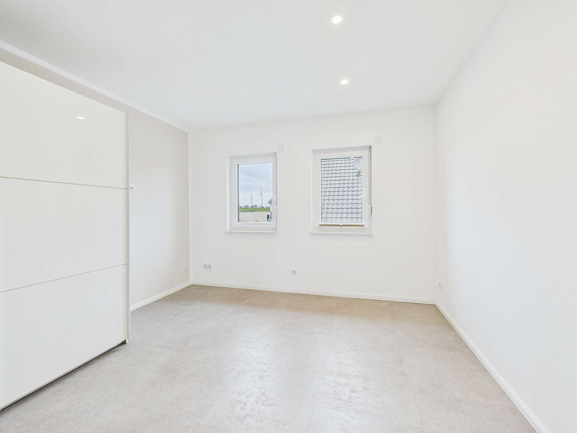 Heller, leerer Raum mit zwei Fenstern und einem weißen Schrank an der Wand. Neutraler, moderner Stil.