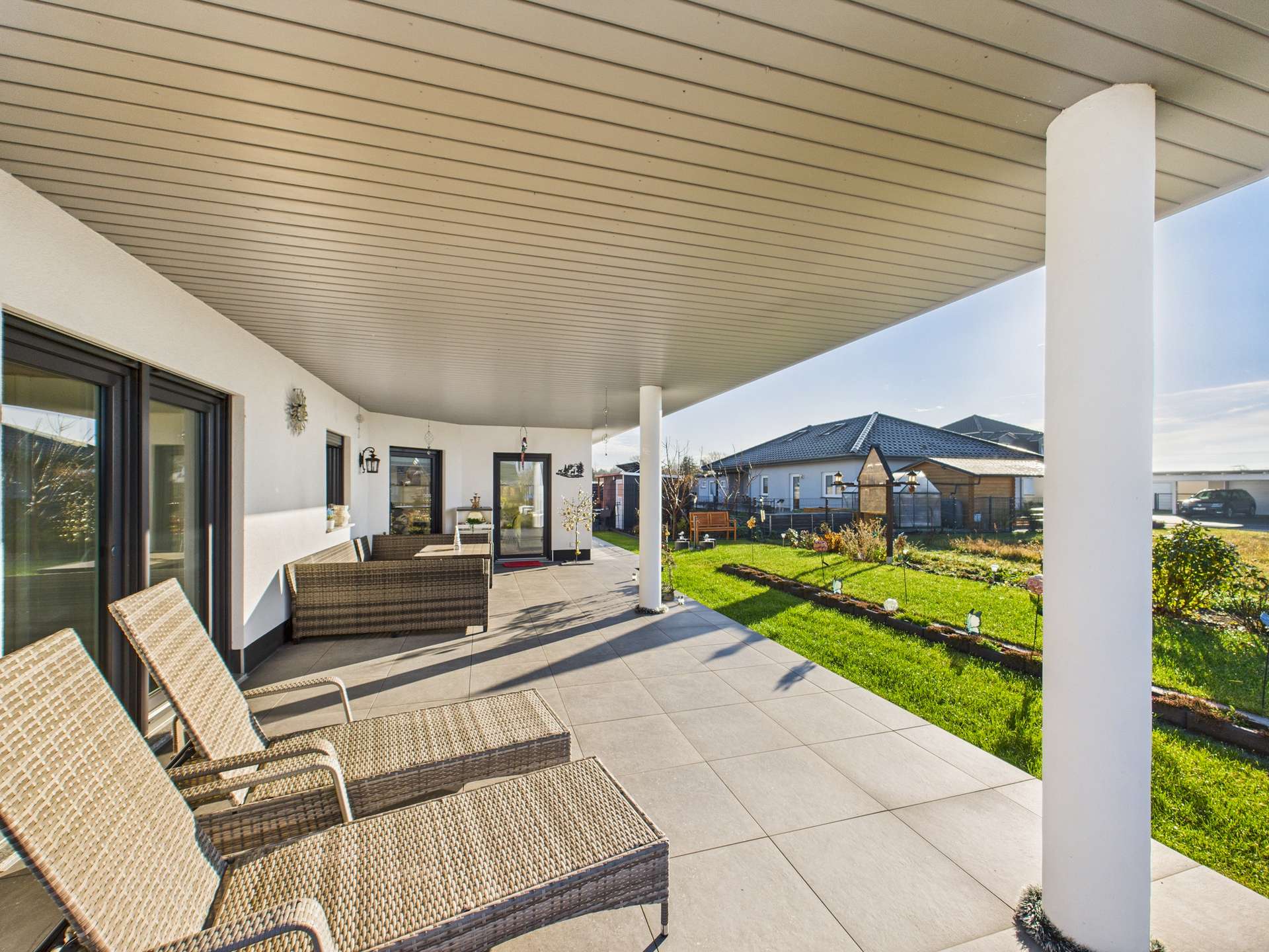 Terrasse mit modernen Liegen, großflächigen Fenstern und Blick auf den Garten bei sonnigem Wetter.