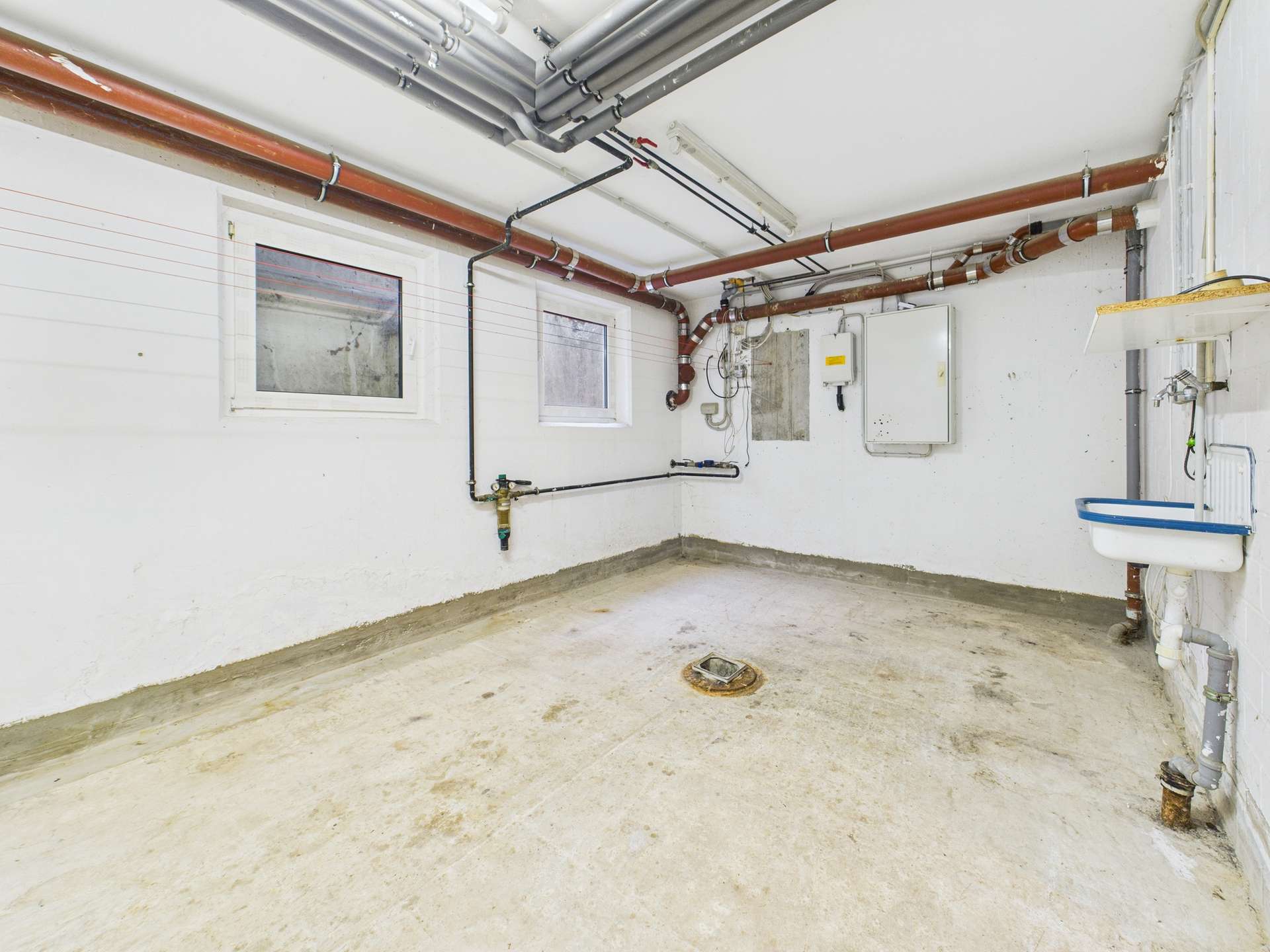 Leerer Keller mit Betonboden, weißen Wänden und sichtbaren Rohren sowie einer Wandhängeschüssel.