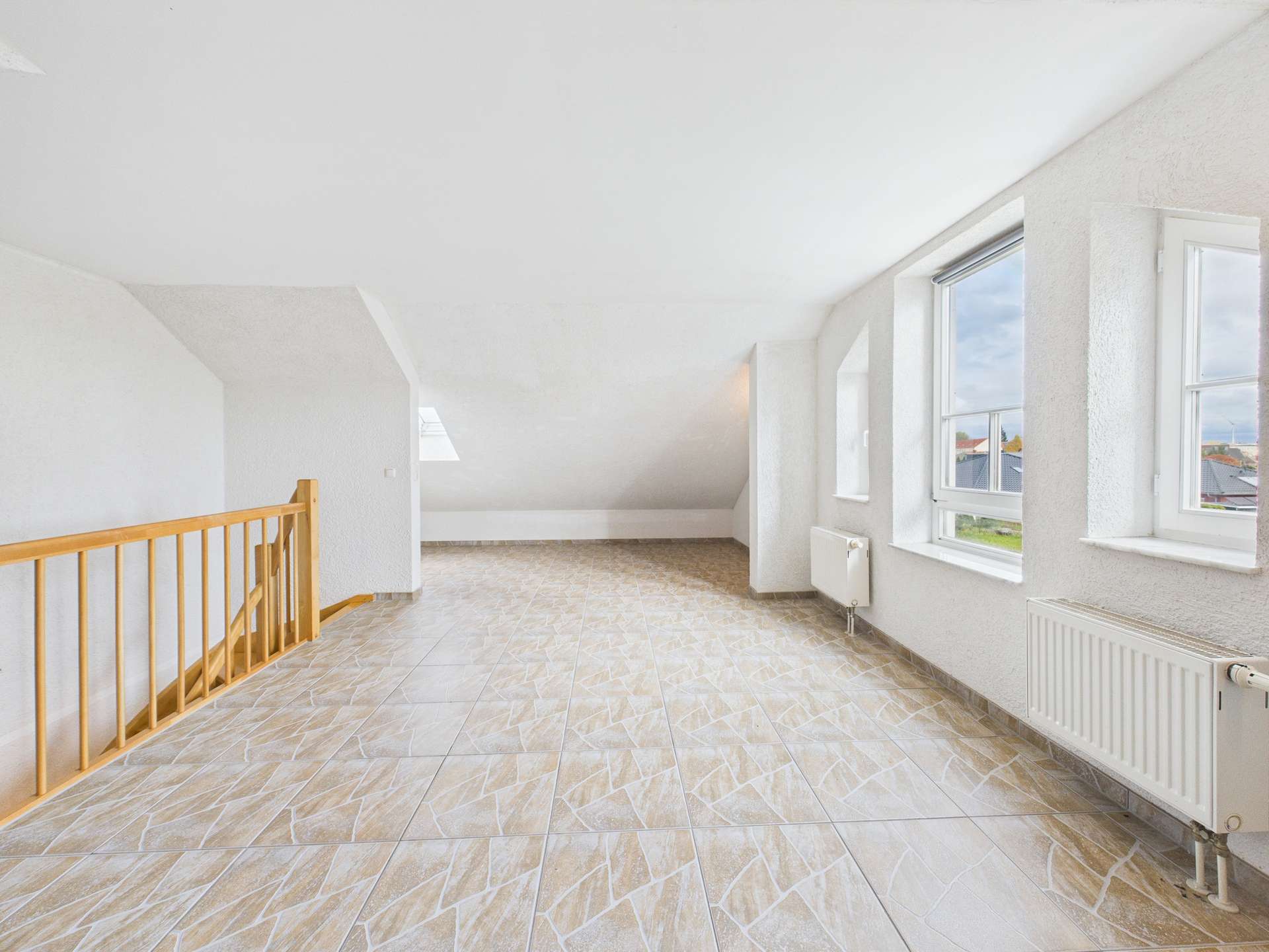 Helles, leeres Zimmer mit schrägen Wänden, großen Fenstern und einem Holzgeländer. Fliesenboden in neutralen Farben.
