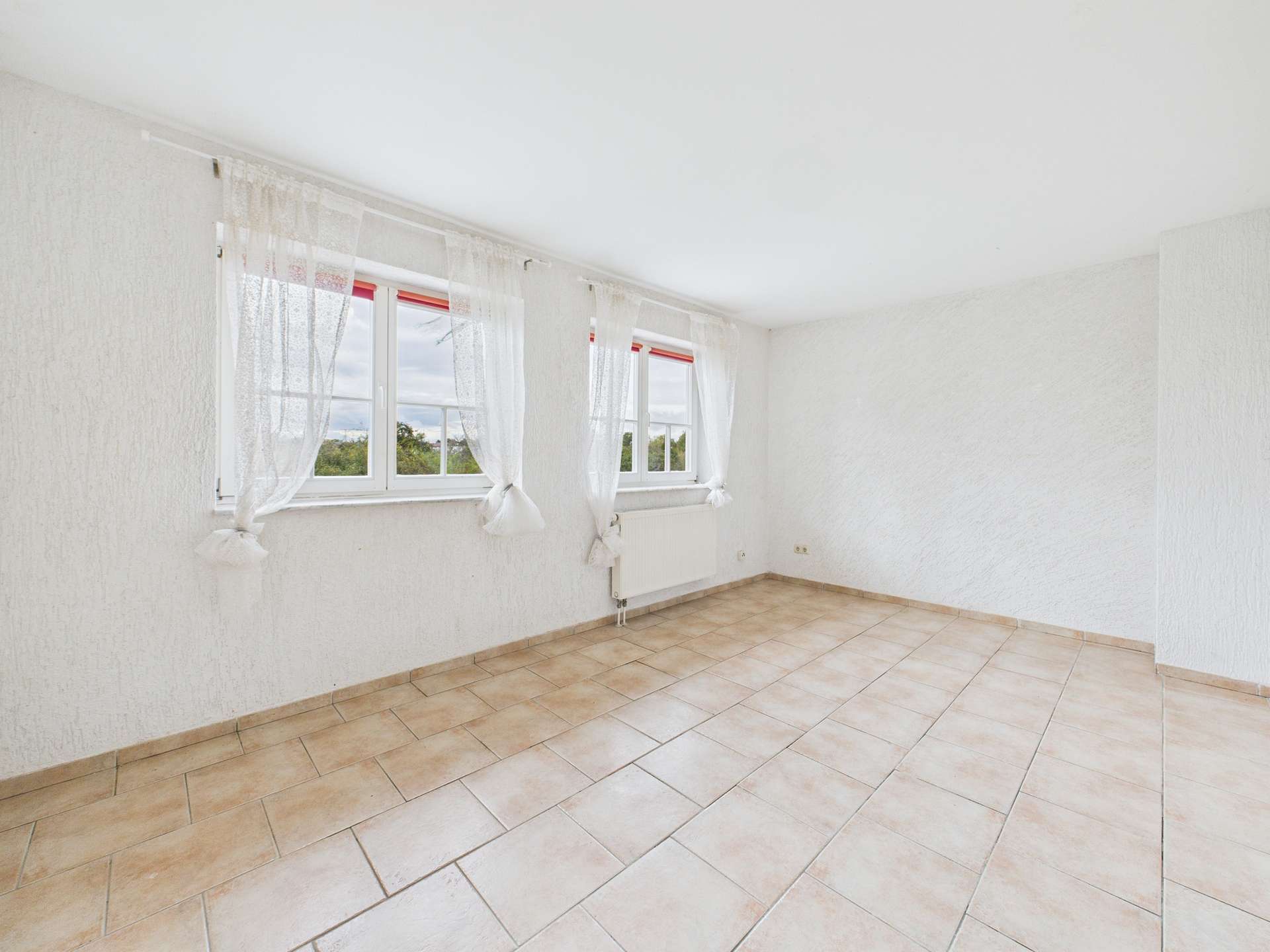 Heller, leerer Raum mit großen Fenstern und Fliesenboden, weiße Wände und Blick auf die Landschaft.