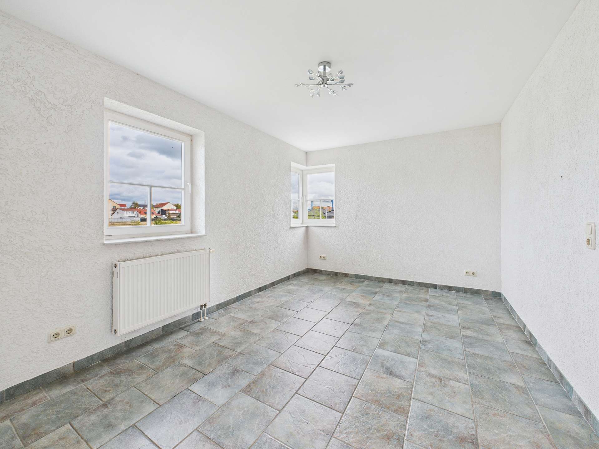 Heller, leerer Raum mit zwei Fenstern, grauen Fliesen und weißem Putz an Wänden. Heizkörper an der Wand.