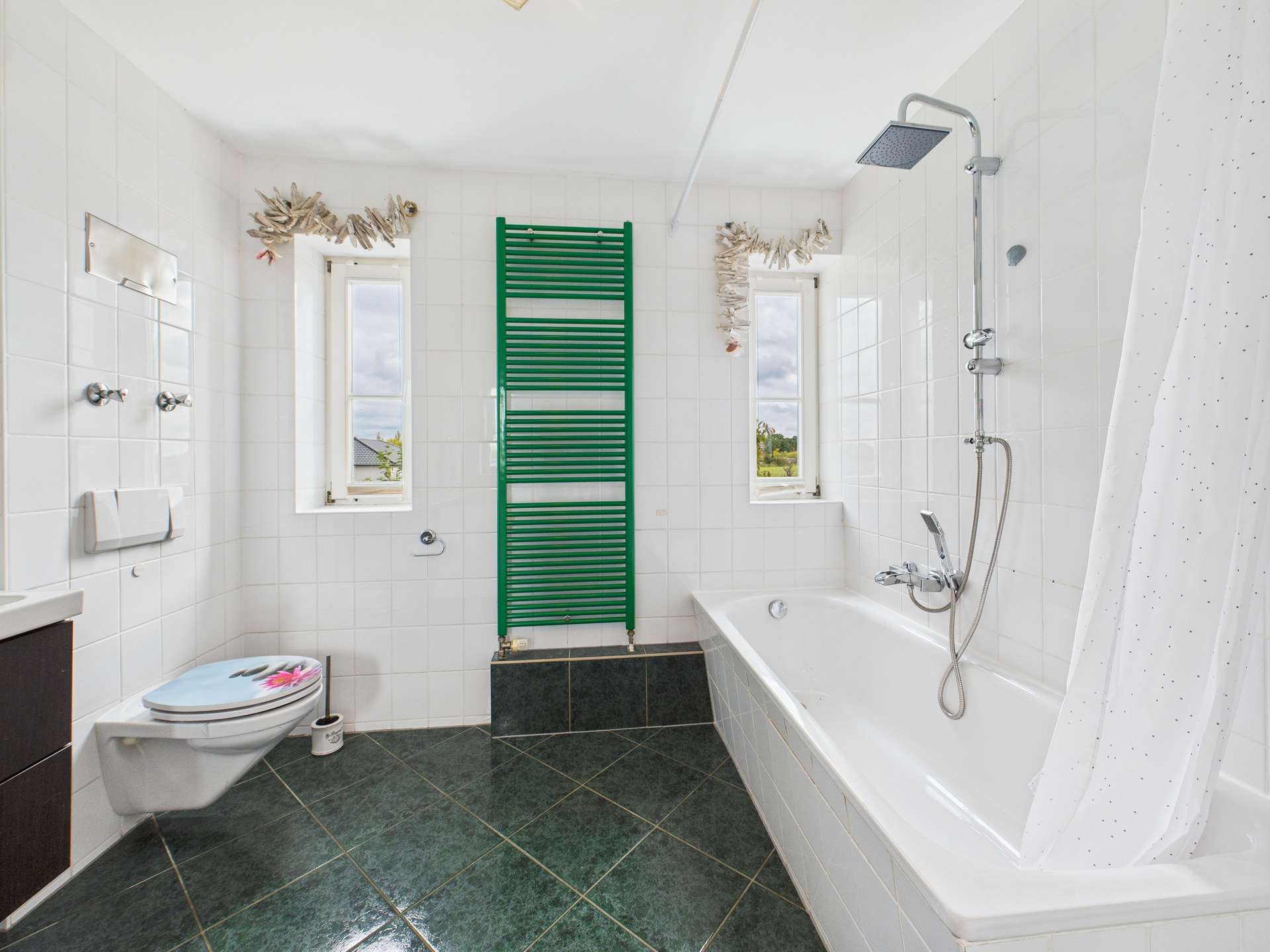 Badezimmer mit Badewanne, Duschkopf, grünem Handtuchhalter und weißen Fliesen. Helle Atmosphäre durch Fenster.